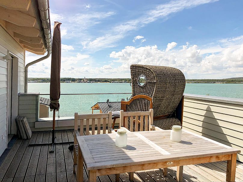 Holz-Dachterrasse mit Esstisch, Strandkorb und eingeklapptem Sonnenschirm. Im Hintergrund der grüne See.